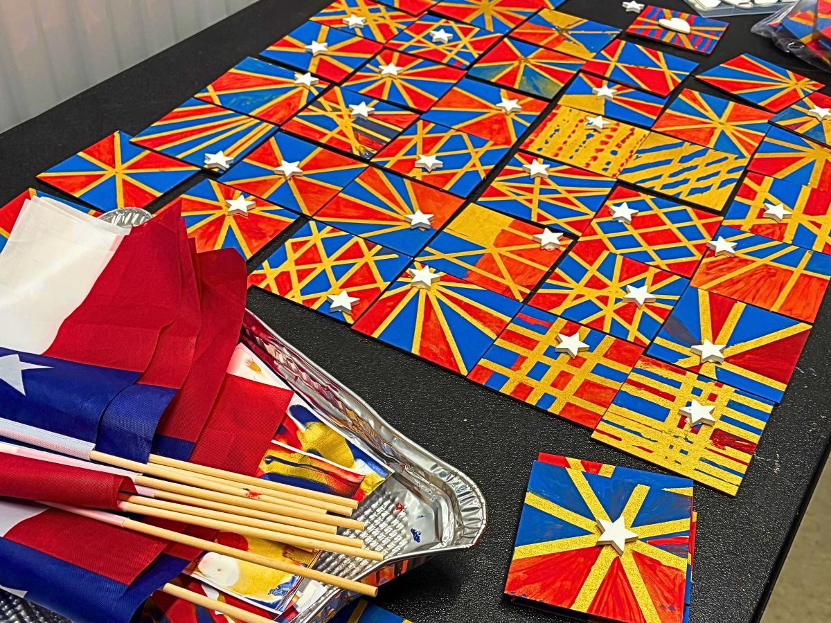 Dozens of hand painted tiles in Texas flag red, white and blue, laid out edge to edge in starburst patterns, with folded Texas flags resting alongside them.
