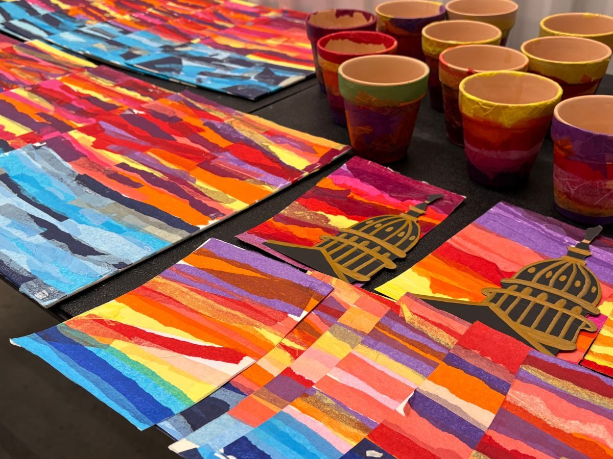 Torn paper collage sunsets in bands of red, orange and violet laid out across a table, several of them cut with a black silhouette of the Capitol dome.
